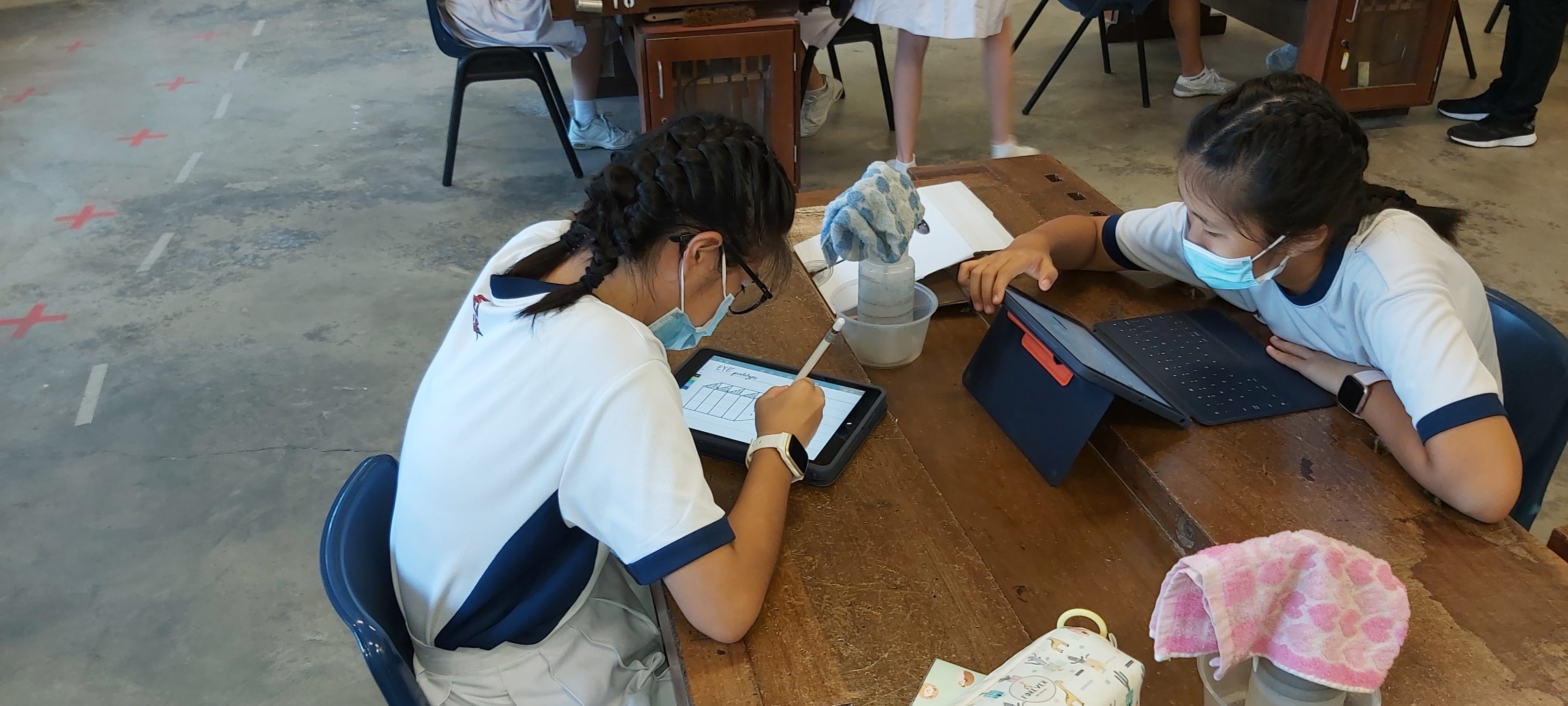 Two masked schoolgirls in uniforms work on tablets at a wooden desk in a classroom.